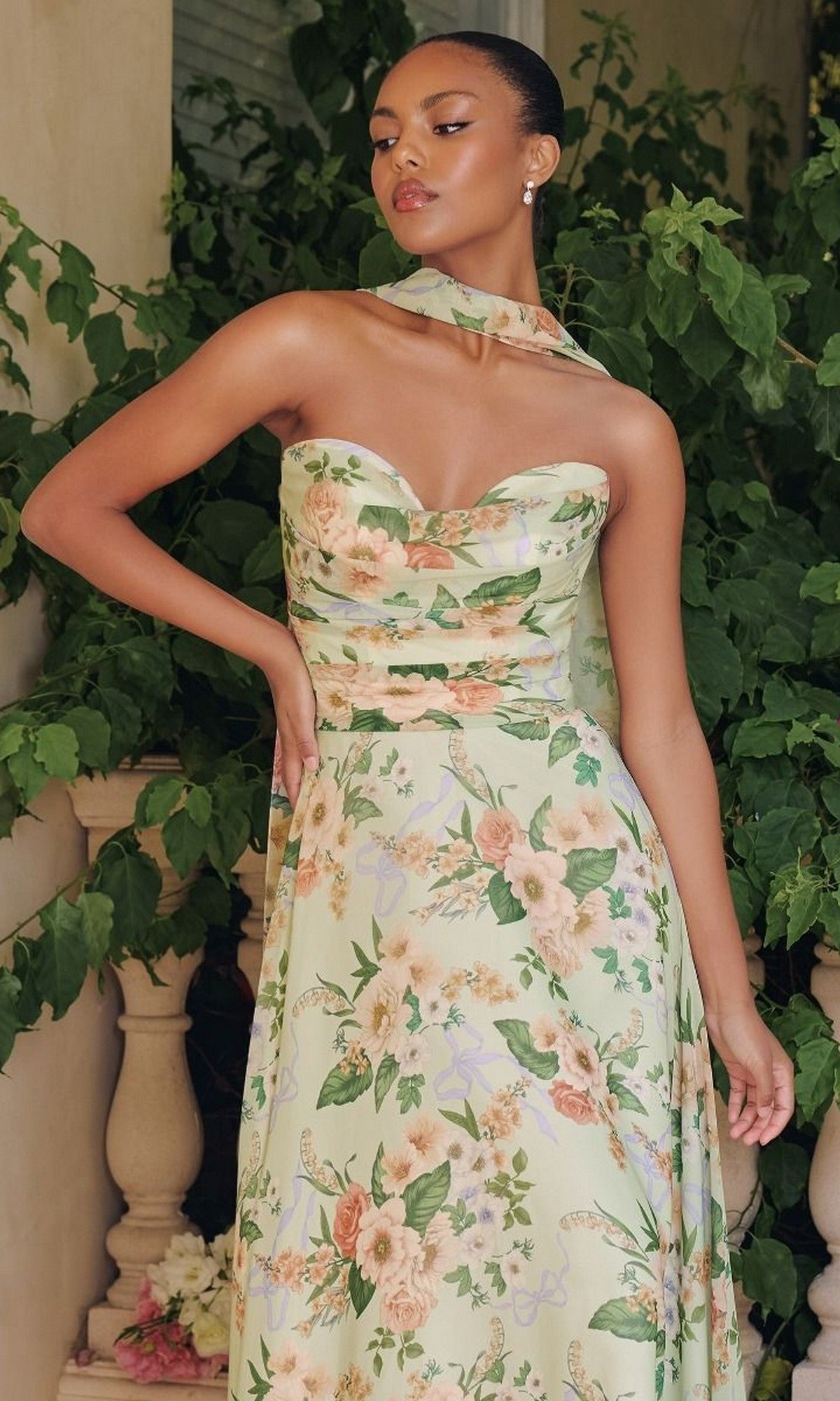 A woman stands before lush greenery, wearing the Amoris T717 strapless long print prom dress in pastel green, paired with a matching sheer scarf draped around her neck.